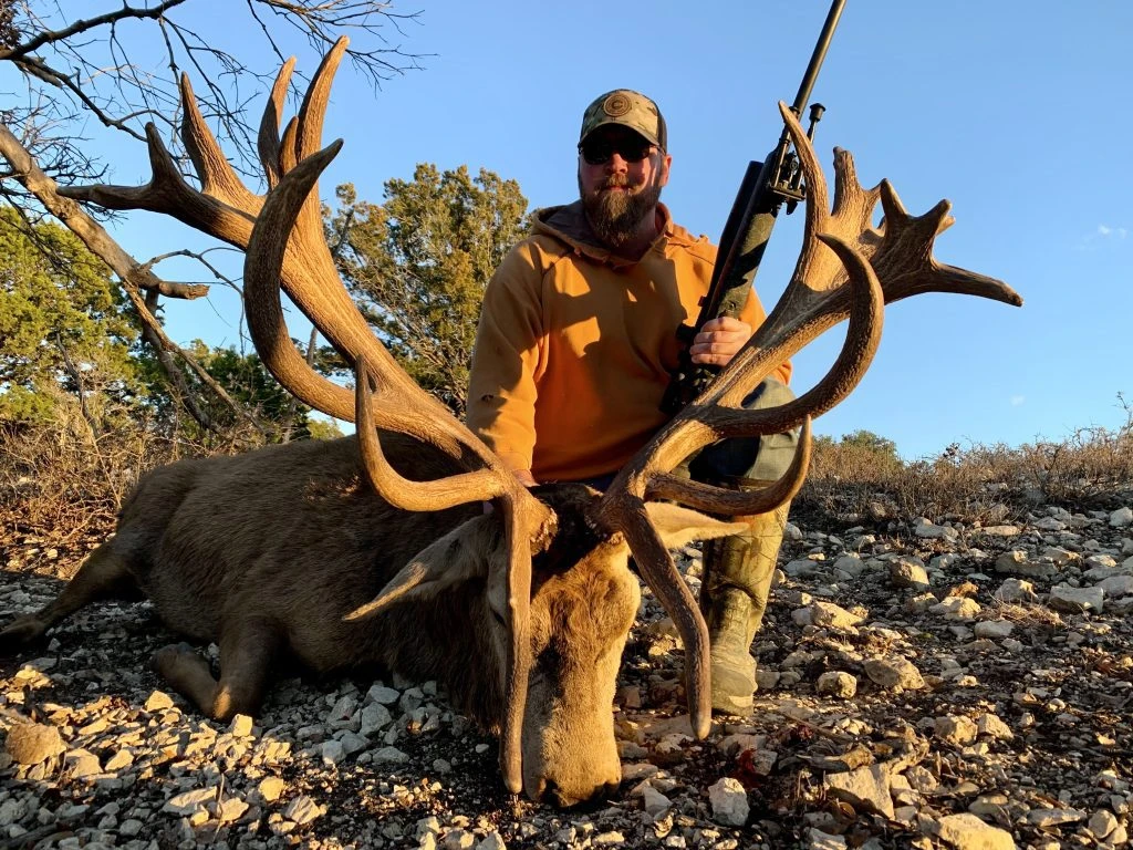 Bearded hunter in orange hoodie posing behind a giant red stag with thick antlers